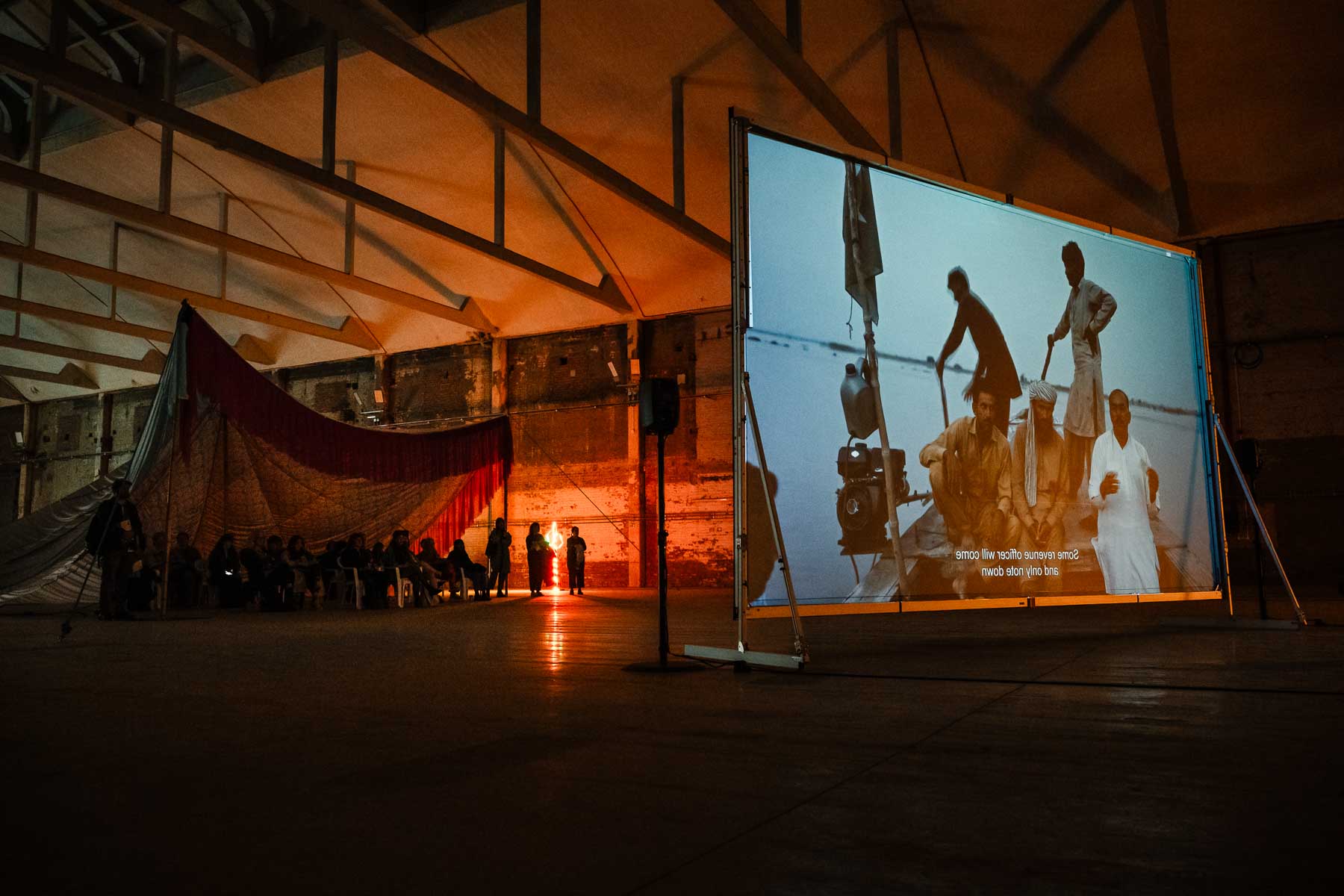 Visitors sit beneath a large fabric canopy watching a film projected onto a freestanding screen in a dark exhibition space.