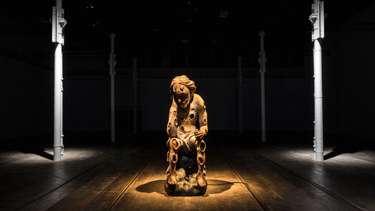 There is a dimly lit room with four thin metal grey columns and wooden floors. We can see a brown sculpted figure in the middle with a warm light spotlight illuminating it from above. The figure is sitting down with their hand holding their head, in a reflexive pose. They have circular carvings all over their body. This is part of an art installation in Tramway, Glasgow International 2018. 