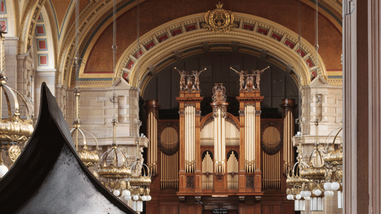 Black conical metal structure with a see-trough hole in it surrounded with a white flowers. The Kelvingrove Museum organ can be seen behind it.