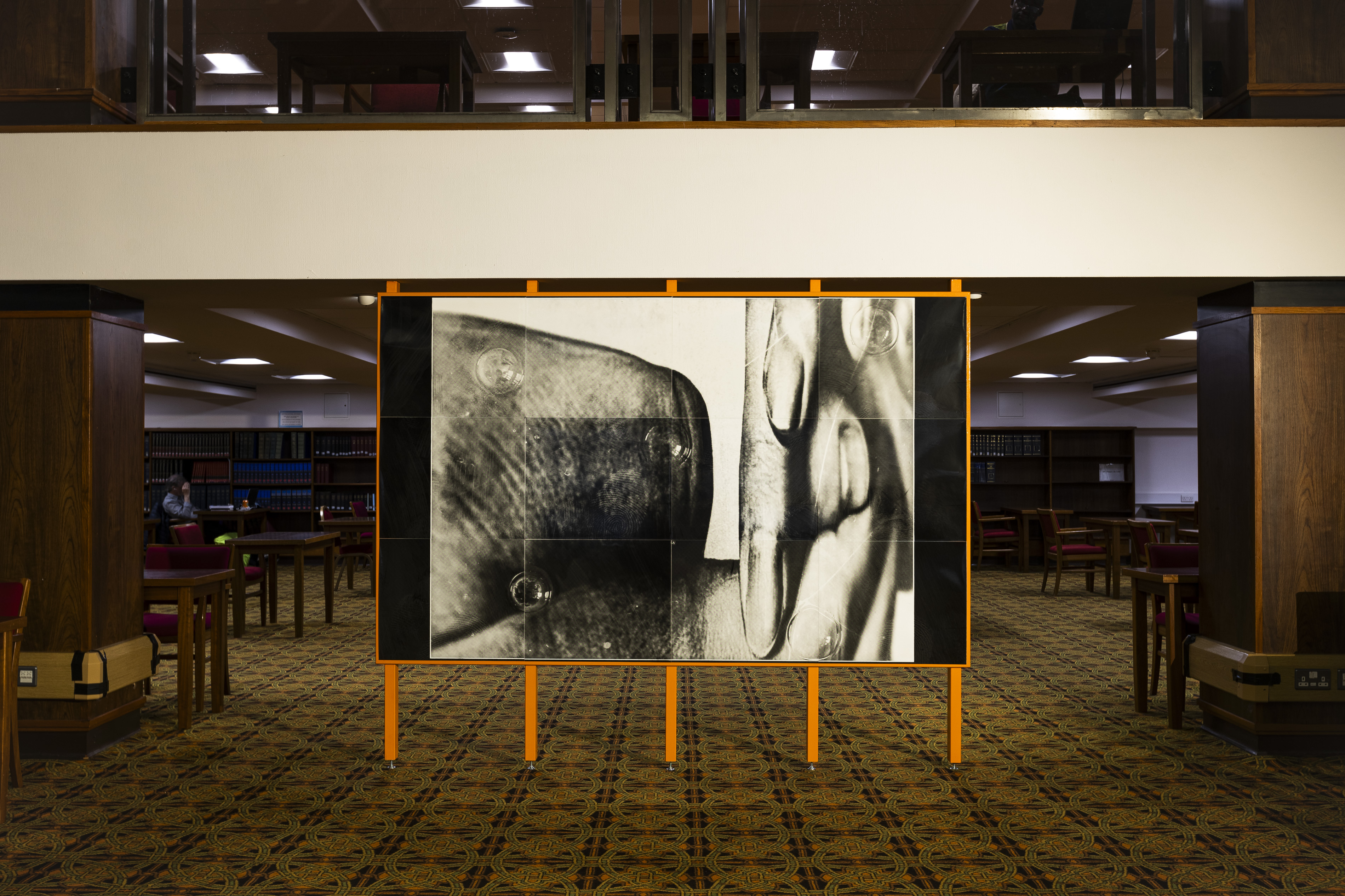 Orange-framed billboard sculpture standing on stilt legs in the Mitchell Library reading room, displaying a black-and-white photographic collage of a hand's fingers beside an eel-like form, with bubbles scattered across the tiled panels. Library shelves and red-upholstered chairs are visible in the background.