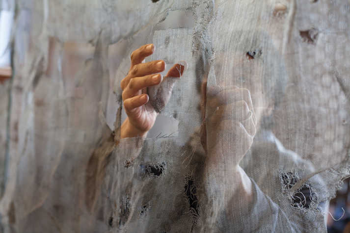 A person is visible behind a screen, holding their hands up to a hole in the screen
