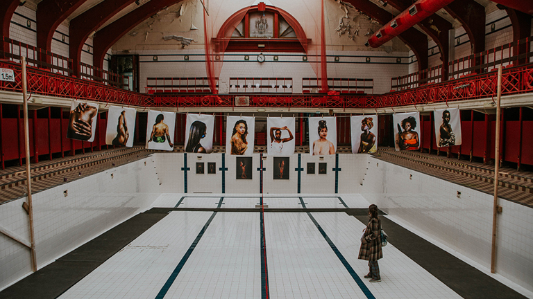 Photographs of women of colour hung across the Govanhill Baths empty swimming pool with festival attendee viewing the work