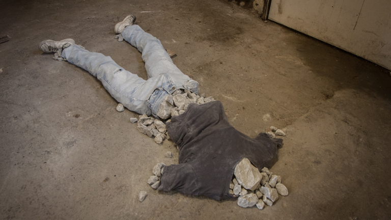 Rough concrete floor with sculptural work. clothes laid out in the shape of a body, with rocks replacing the body