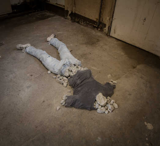 Rough concrete floor with sculptural work. clothes laid out in the shape of a body, with rocks replacing the body