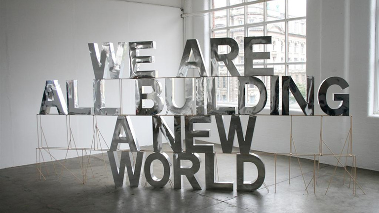 sculpture of letters in a gallery space reading, 'we are all building a new world'