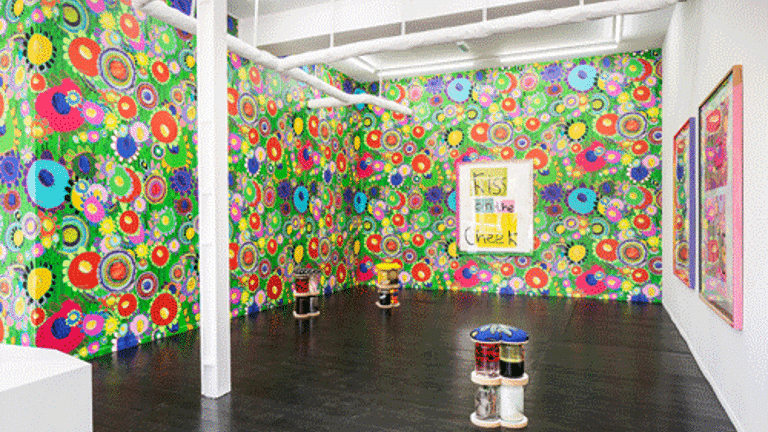 The image shows a colourful exhibition with bright patterned wallpaper and sculptures made of glass jars