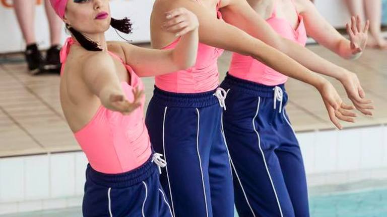 Three figures wearing pink swimming caps, pink swimming suits, and blue tracksuit legs perform next to a swimming pool