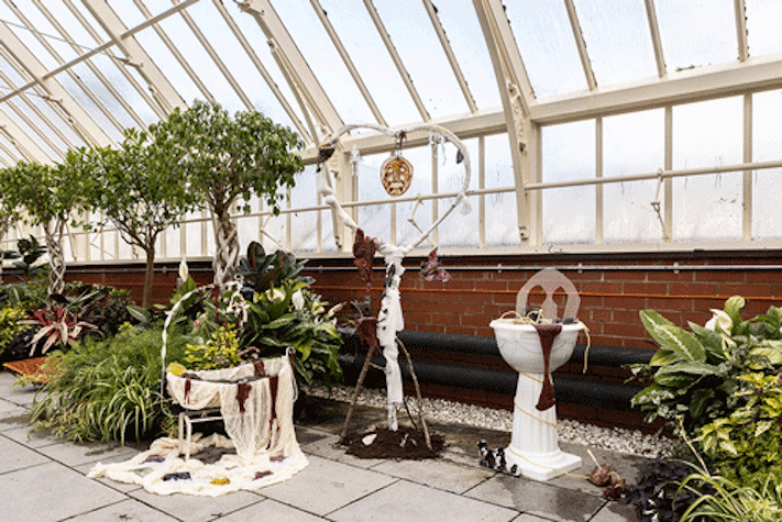 An interior glasshouse room filled with a variety of plants and trees, with a small sculptures are positioned in front of plants.