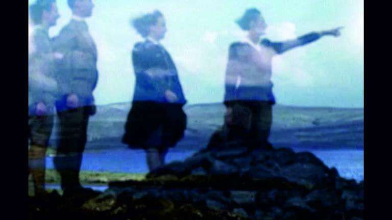 Film still showing four overlaid figures against a background of a lake and mountains. One figure points to the right, toward something outside the frame.
