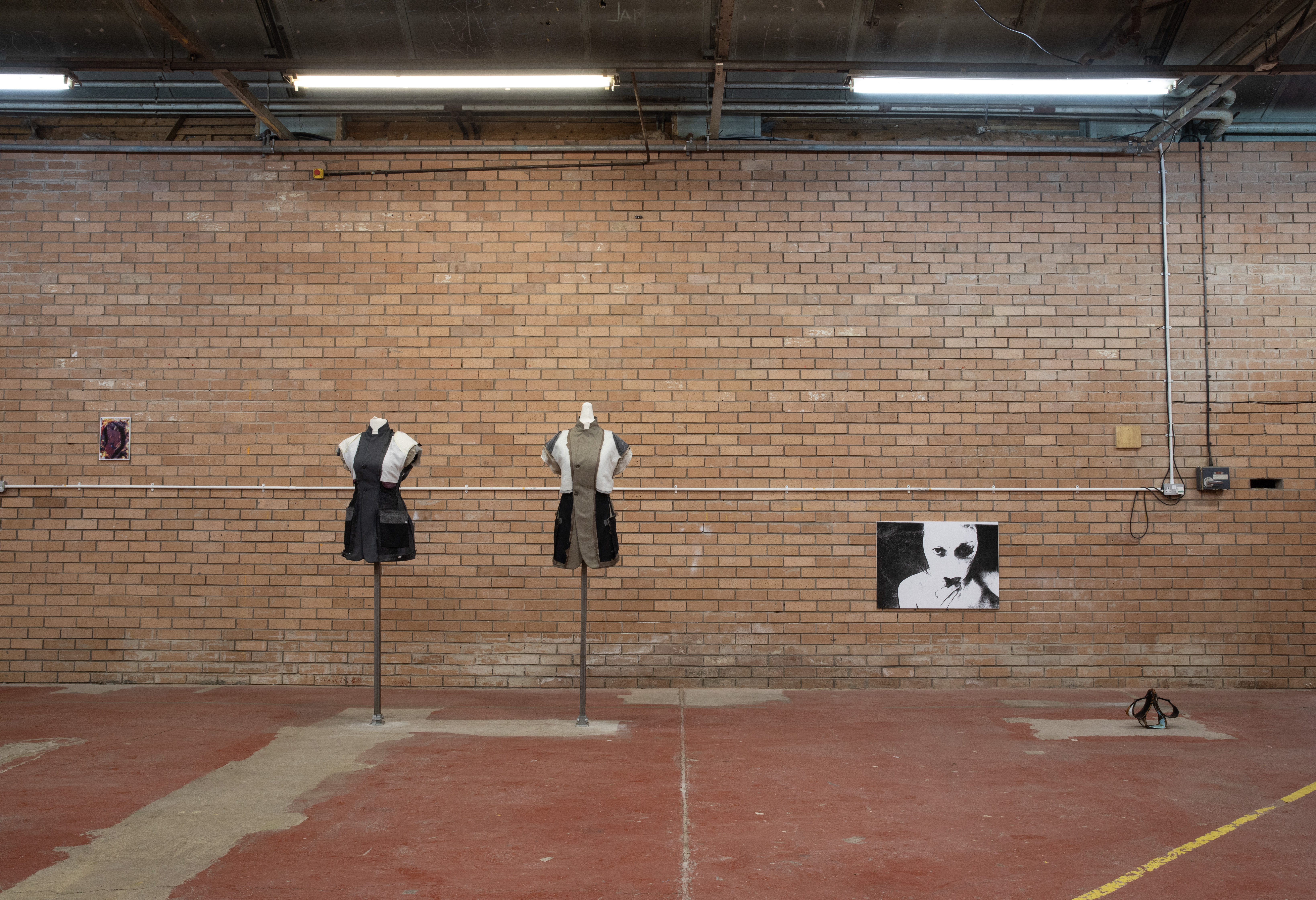 Two headless, sleeveless garments in black and khaki stand on poles against a brick wall, flanked by a small colour print and a black and white portrait.