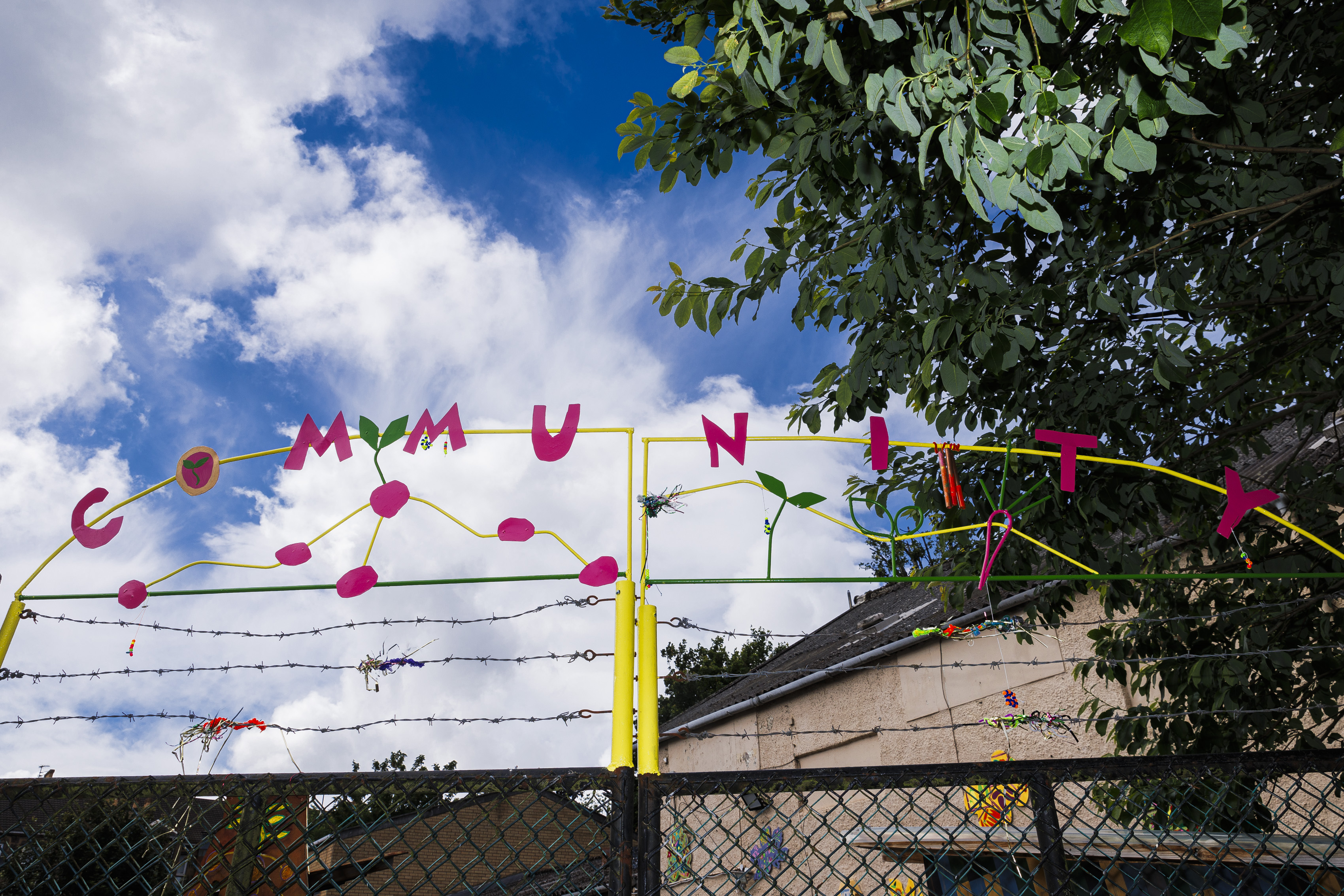 Community. Photograph of a metal sign above a gate reading "Community," decorated with a small metal potato shapes growing around the letters. The metalwork is painted pink, green, and yellow and appears against a bright blue sky and a tree in full blood. 