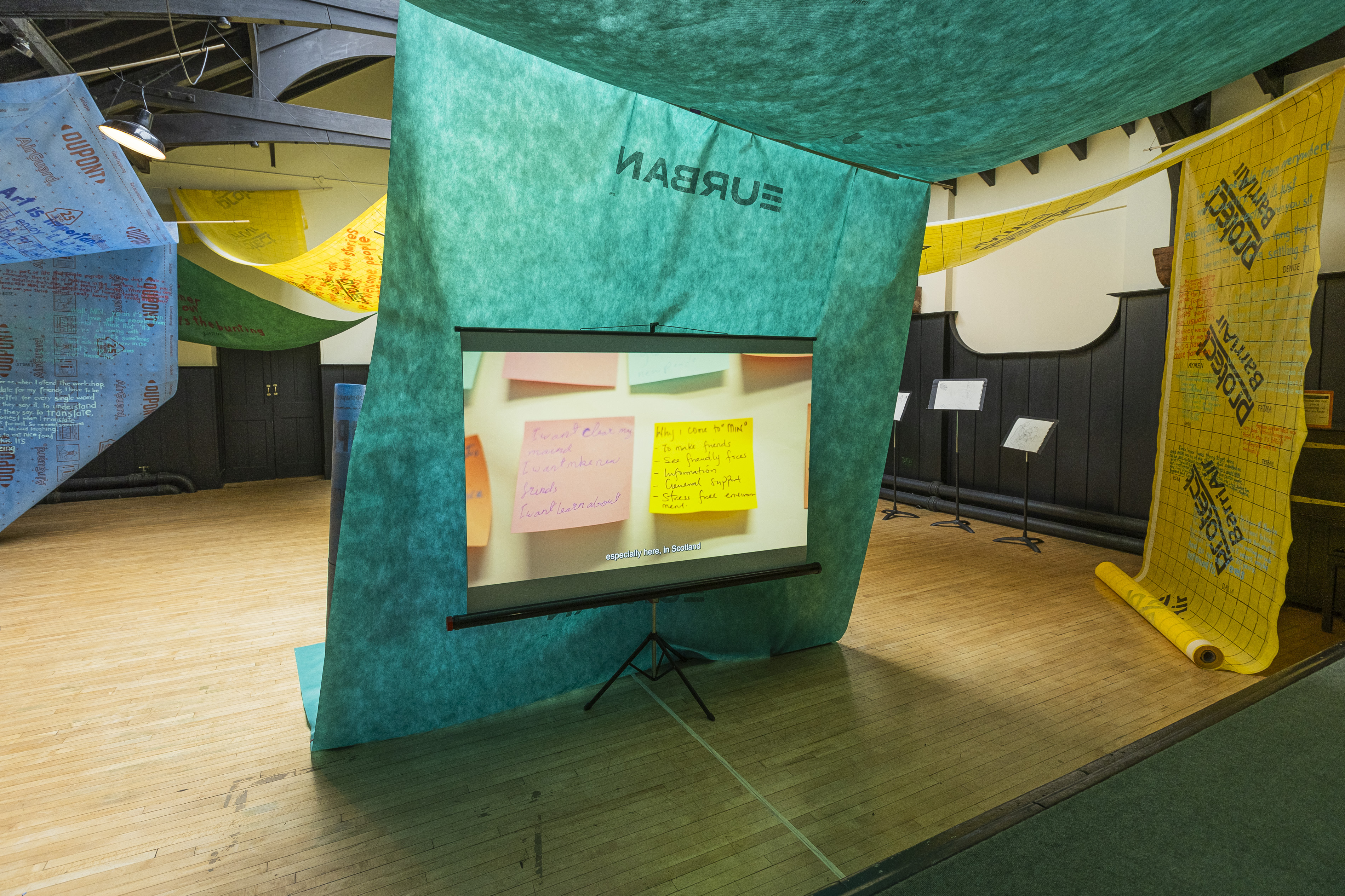 Interior of a tent-like installation made from a green fabric banner reading "EURBAN" (reversed), housing a projector screen displaying handwritten notes on coloured paper with the subtitle "especially here, in Scotland", surrounded by hanging yellow and blue printed banners and music stands holding sketchbooks.