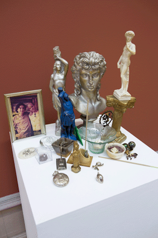 A short plinth is stood against the wall. It has multiple sculptures and small objects placed on it. There is a bust of a man with long curly hair, two sculptures of men in different poses, as one is tied to a wooden pole. There is a vase, a pocket watch, a picture frame with the picture of to men standing next to each other among them.