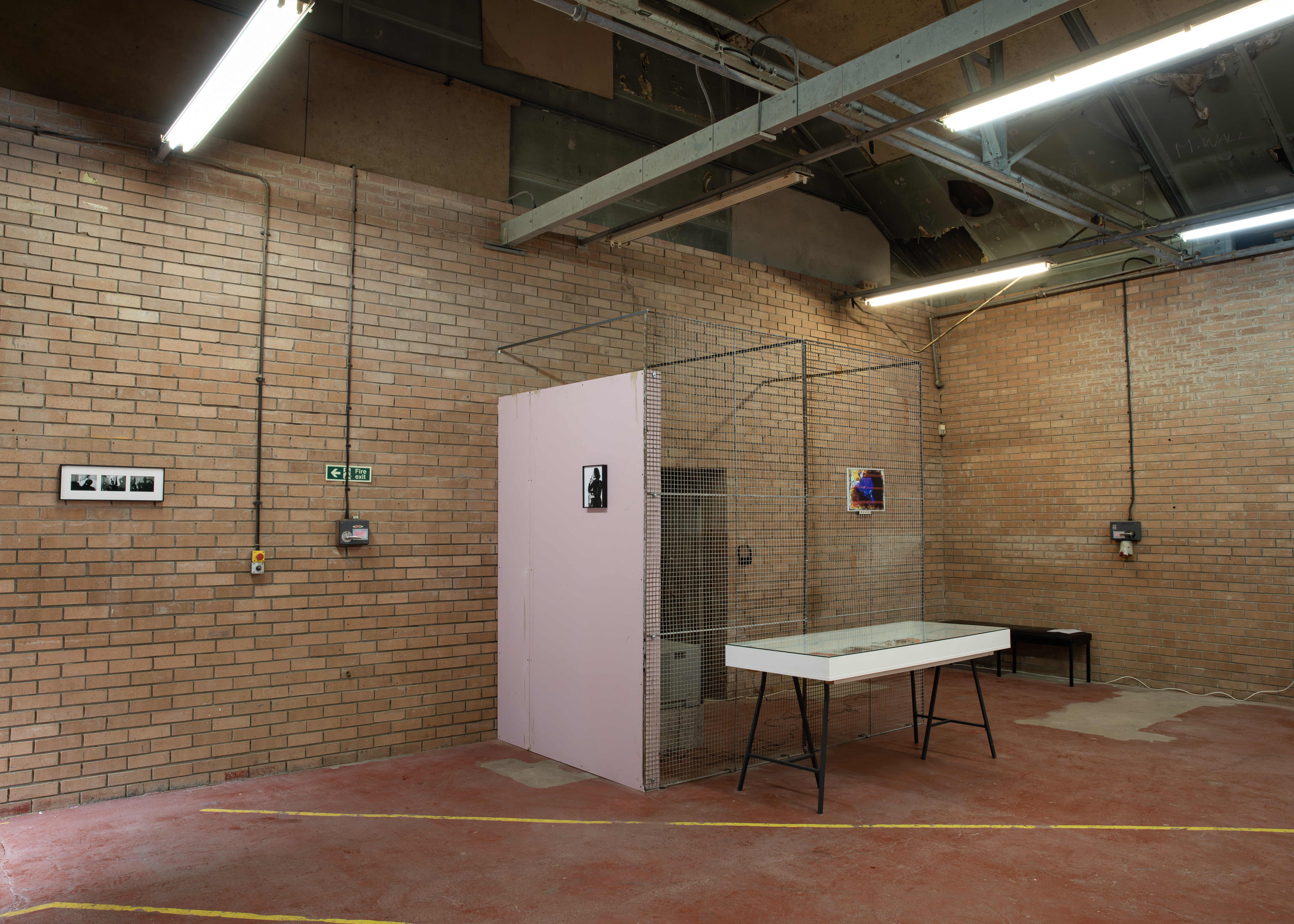 A pink partition wall and wire-mesh cage structure with framed photographs, alongside a glass-topped display table on trestle legs in a brick-walled space.