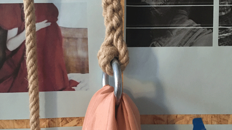 Close up photo of a rope with metal ring and a section of pink material hanging from it. This is in front of a board with images stuck on it