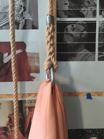 Close up photo of a rope with metal ring and a section of pink material hanging from it. This is in front of a board with images stuck on it