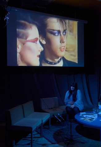 A screening of 'The Height of Goth: 1984, A Night at the Xclusiv Nightclub, Batley, West Yorkshire'. Below, Claire Biddles sitting on a couch, under blue light.