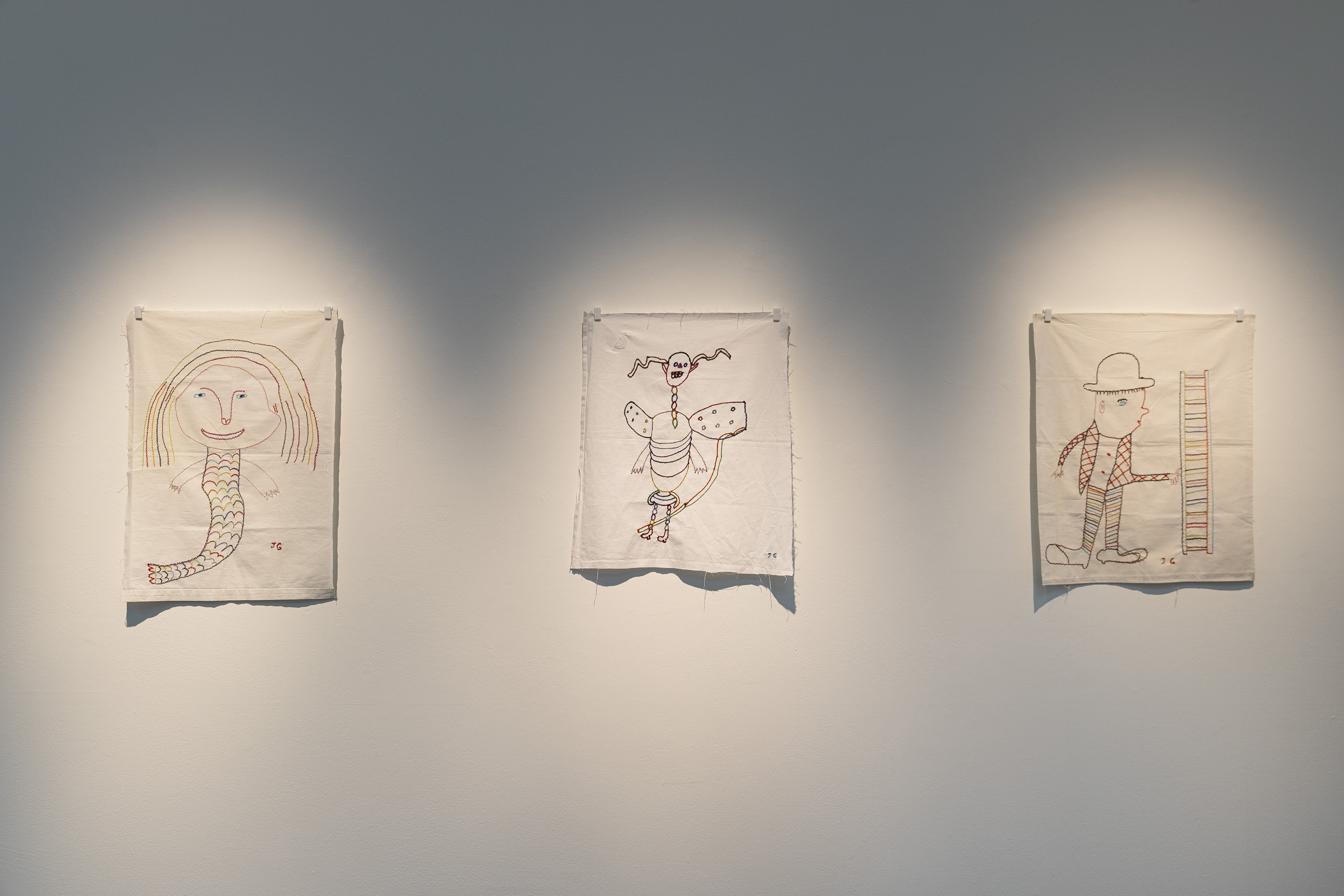 Three embroidered fabric works by James Gladwell hung side by side, depicting a mermaid-like figure, a winged creature with a long tail, and a man in a hat and striped trousers standing beside a ladder.