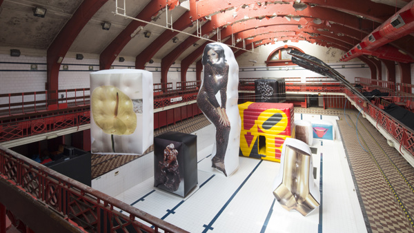 In an empty swimming pool. There are eight artworks, printed on inflated boxes. A sculpture of two men kissing. On its left is a large inflated box with the image of a flower. In the centre is a woman in a leopard jumpsuit printed on a giant inflated piece in the same shape. Behind it, multicoloured cube with the words LOVE. On the left, the shape of a wrench. Behind it, a sculpture of two women in an embrace. Lastly, one with a picture of a mouth with a tongue sticking out.