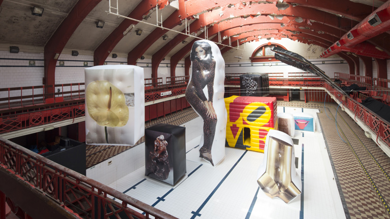 In an empty swimming pool. There are eight artworks, printed on inflated boxes. A sculpture of two men kissing. On its left is a large inflated box with the image of a flower. In the centre is a woman in a leopard jumpsuit printed on a giant inflated piece in the same shape. Behind it, multicoloured cube with the words LOVE. On the left, the shape of a wrench. Behind it, a sculpture of two women in an embrace. Lastly, one with a picture of a mouth with a tongue sticking out.