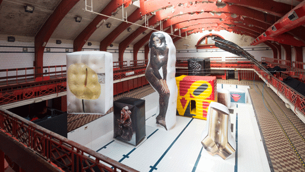 In an empty swimming pool. There are eight artworks, printed on inflated boxes. A sculpture of two men kissing. On its left is a large inflated box with the image of a flower. In the centre is a woman in a leopard jumpsuit printed on a giant inflated piece in the same shape. Behind it, multicoloured cube with the words LOVE. On the left, the shape of a wrench. Behind it, a sculpture of two women in an embrace. Lastly, one with a picture of a mouth with a tongue sticking out.