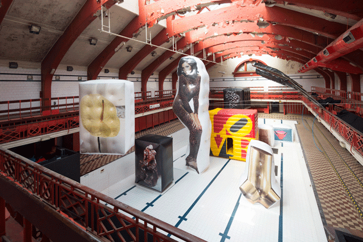 In an empty swimming pool. There are eight artworks, printed on inflated boxes. A sculpture of two men kissing. On its left is a large inflated box with the image of a flower. In the centre is a woman in a leopard jumpsuit printed on a giant inflated piece in the same shape. Behind it, multicoloured cube with the words LOVE. On the left, the shape of a wrench. Behind it, a sculpture of two women in an embrace. Lastly, one with a picture of a mouth with a tongue sticking out.
