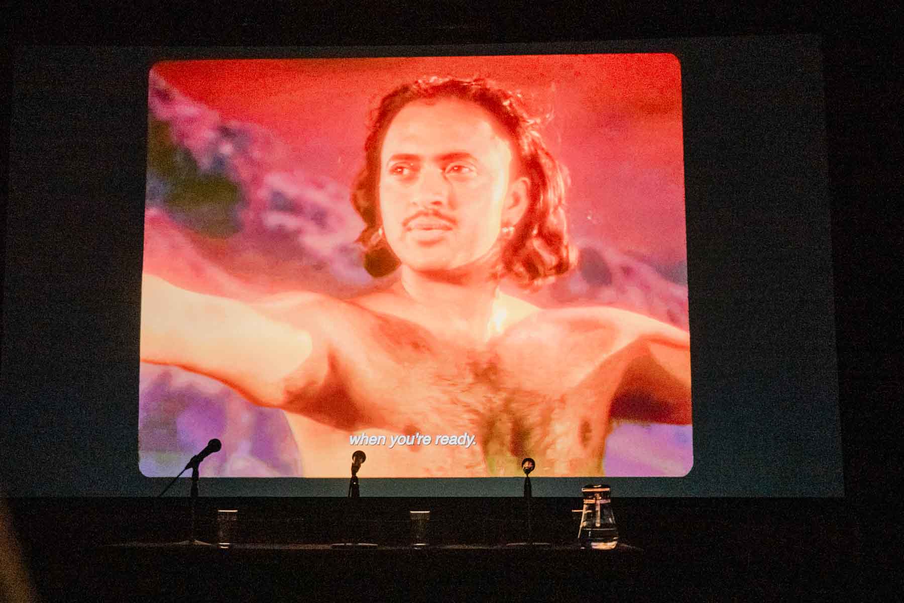 A film still projected on a cinema screen shows a shirtless man with shoulder-length hair standing with his arms outstretched against a pink-toned sky, with the subtitle "when you're ready" visible at the bottom. Microphones and glasses are set up on a panel table in front of the screen.