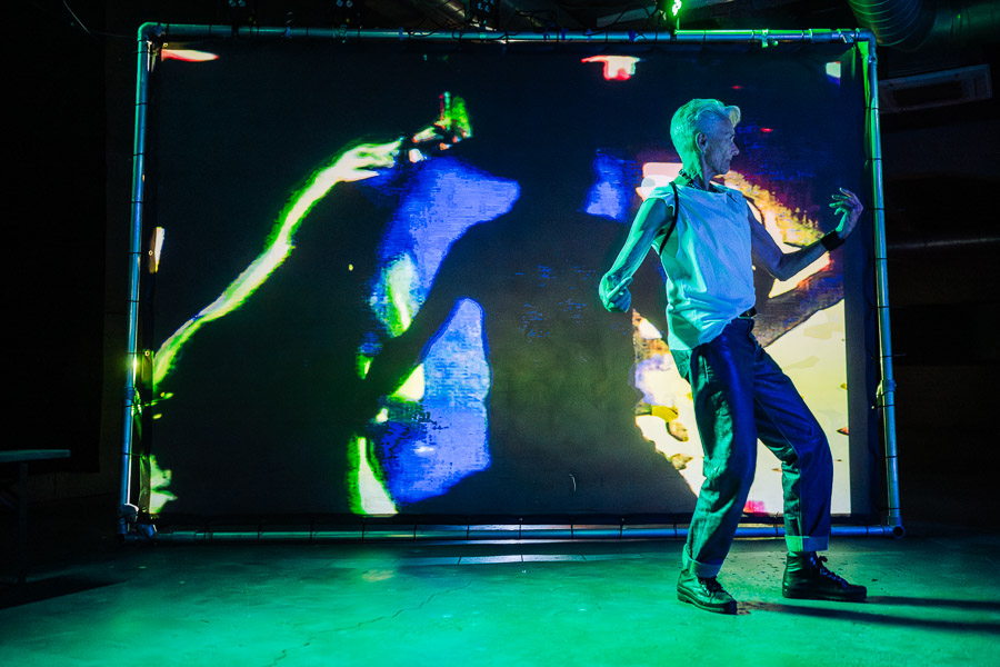An older performer with cropped white hair, wearing a white top and dark trousers, dances under green lighting in front of a screen showing an abstract glitched projection.