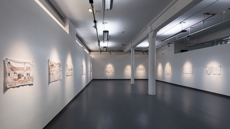 A gallery corridor lined with James Gladwell's embroidered fabric works, each depicting simple, colourful figures and buildings on white cloth, hung along both walls under track lighting.