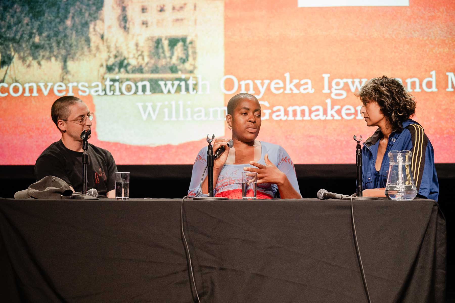 Three people sit at a panel table on a stage, two speaking to each other, in front of a large screen reading "In-conversation with Onyeka Igwe and Michelle Williams Gamaker" alongside the word "Gatherings" and a Glasgow International festival logo.