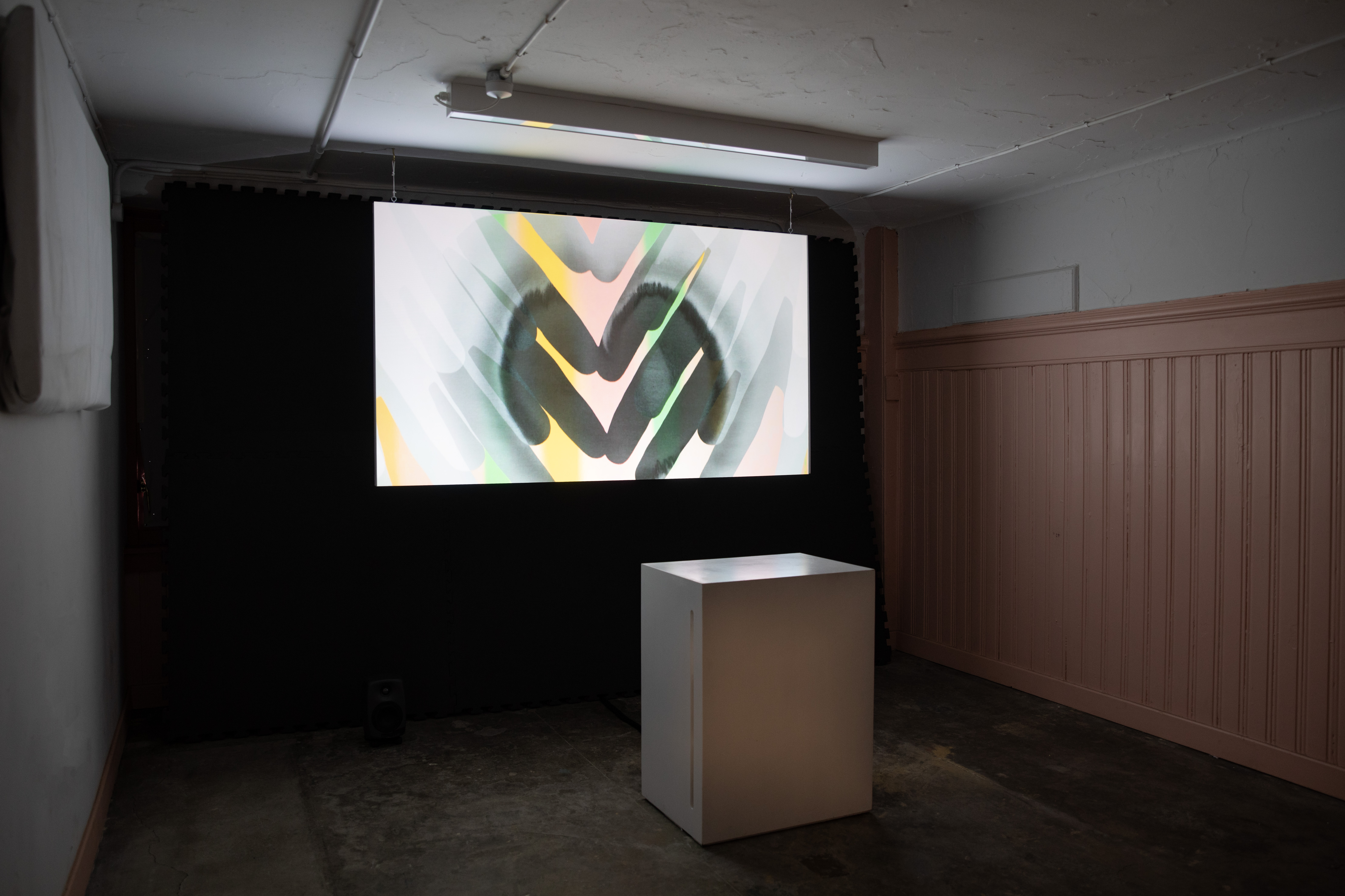 Darkened gallery space with a wide screen showing an abstract video work in soft pastel tones with a dark circular motif at its centre. A white plinth stands beneath the screen, against a wall lined with pink wood panelling.