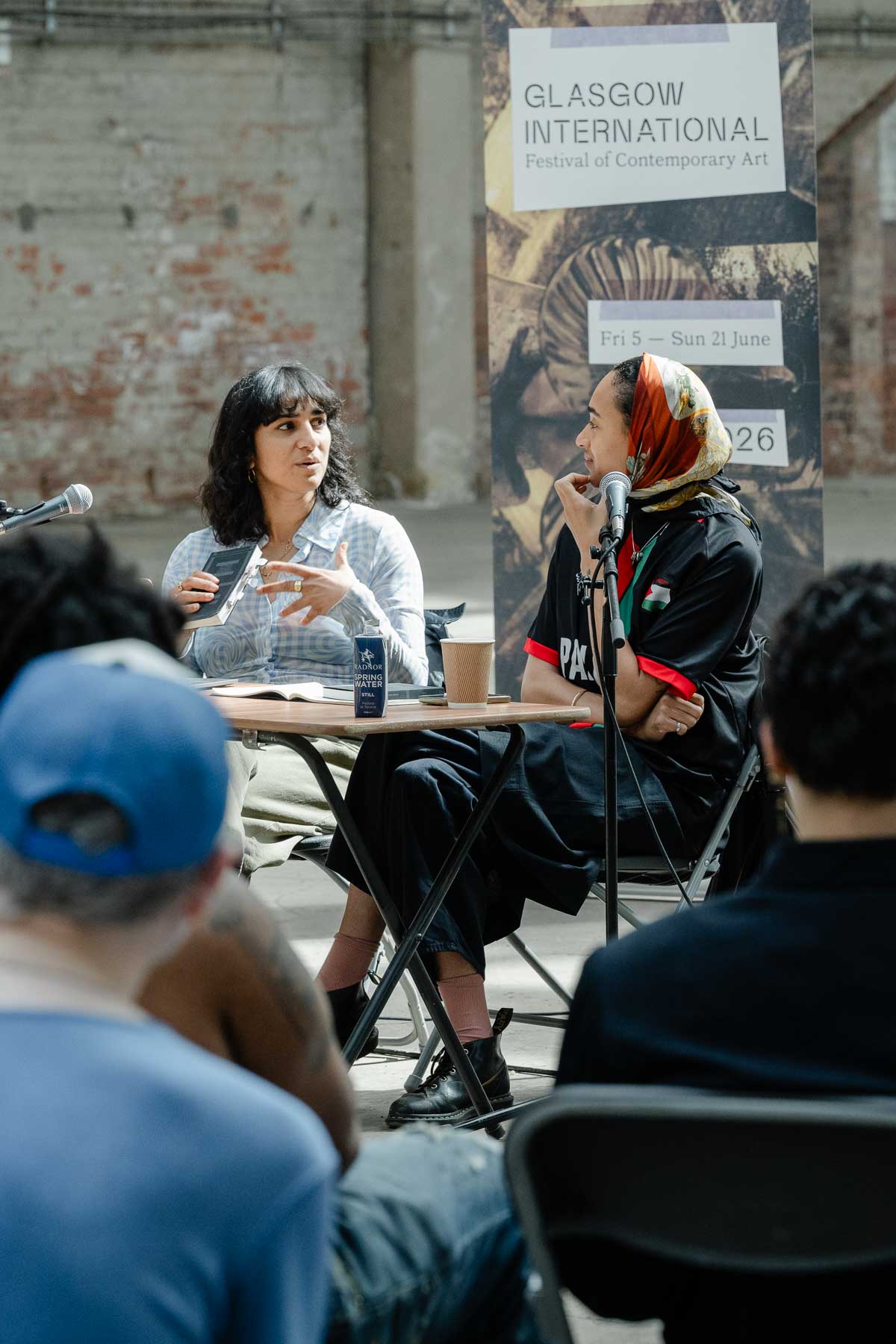 Two women in conversation at a small table, seen from behind an audience. One speaks with her hand raised while holding a book; the other, wearing a headscarf and "Palestine" football shirt, rests her chin on her hand. A Glasgow International banner is behind them.