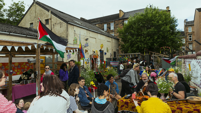 Summer Solstice Celebration. Photograph of people gathered in the yard at Rumpus Room. Some talk while others play intstruments, and drinks are served under a shelter with a Palestine flag flying above it. 