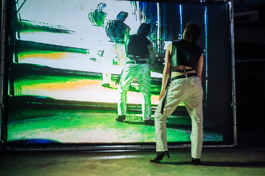 A performer in a leather waistcoat and cream trousers stands with their back to camera, facing a large screen showing a distorted, glitching green-toned projection of two embracing figures.