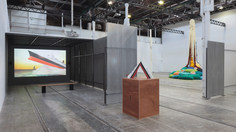 Installation view of a video showing a large boat grounded on a large rock, and tipped up out of the water. Also in view are other sculptures.