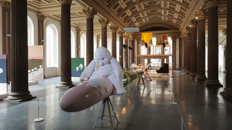 A gallery with a high vaulted ceiling and columns. In the middle a large, white toy-like cartoon figure sits on what appears to be rocket (sculpture). Other art works are visible in the background