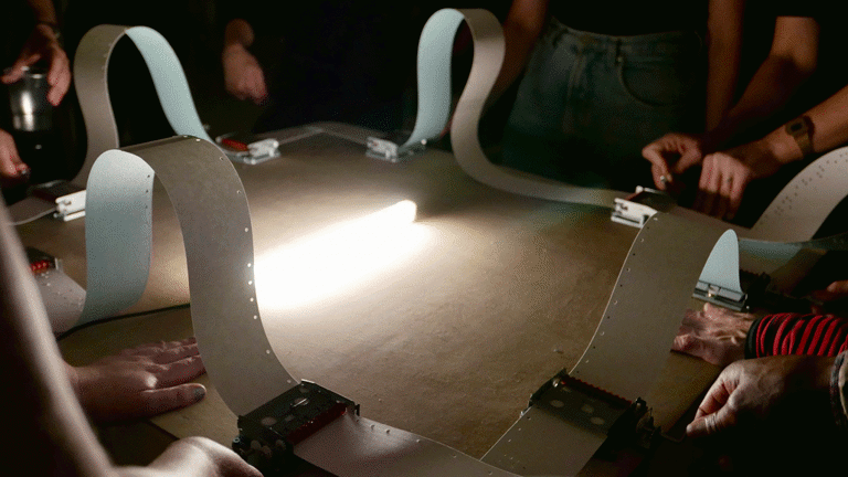 A photograph of multiple sets of hands operating some equipment with tape, and a bright light shining in the middle 