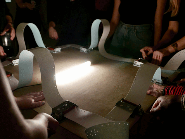 A photograph of multiple sets of hands operating some equipment with tape, and a bright light shining in the middle