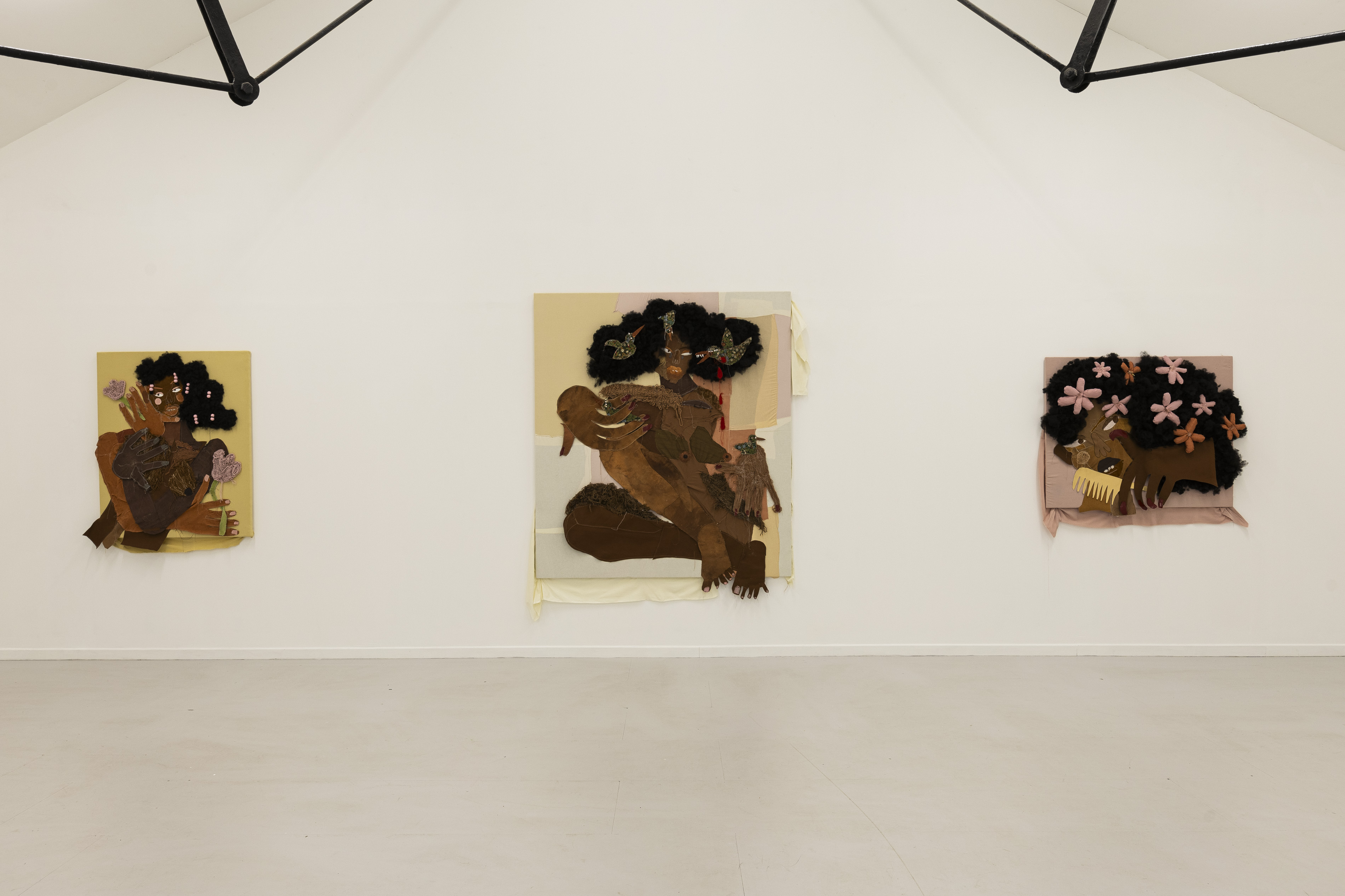 Gallery installation view of three mixed-media textile portraits by Anya Paintsil, each depicting figures with sculptural black woollen hair, oversized fabric hands and exaggerated features, hung across a white wall.