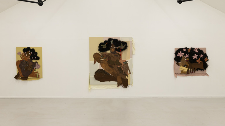 Gallery installation view of three mixed-media textile portraits by Anya Paintsil, each depicting figures with sculptural black woollen hair, oversized fabric hands and exaggerated features, hung across a white wall.