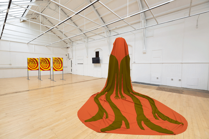 a cape with a long train is suspended from the ceiling of a large room and draped across the floor. On the orange fabric a khaki fabric appliqué depicts two figures back to back, their long legs stretching down the train and ending in heeled shoes. In the background three metal frames hold identical poster images of a red and yellow swirl with black text overlaid.