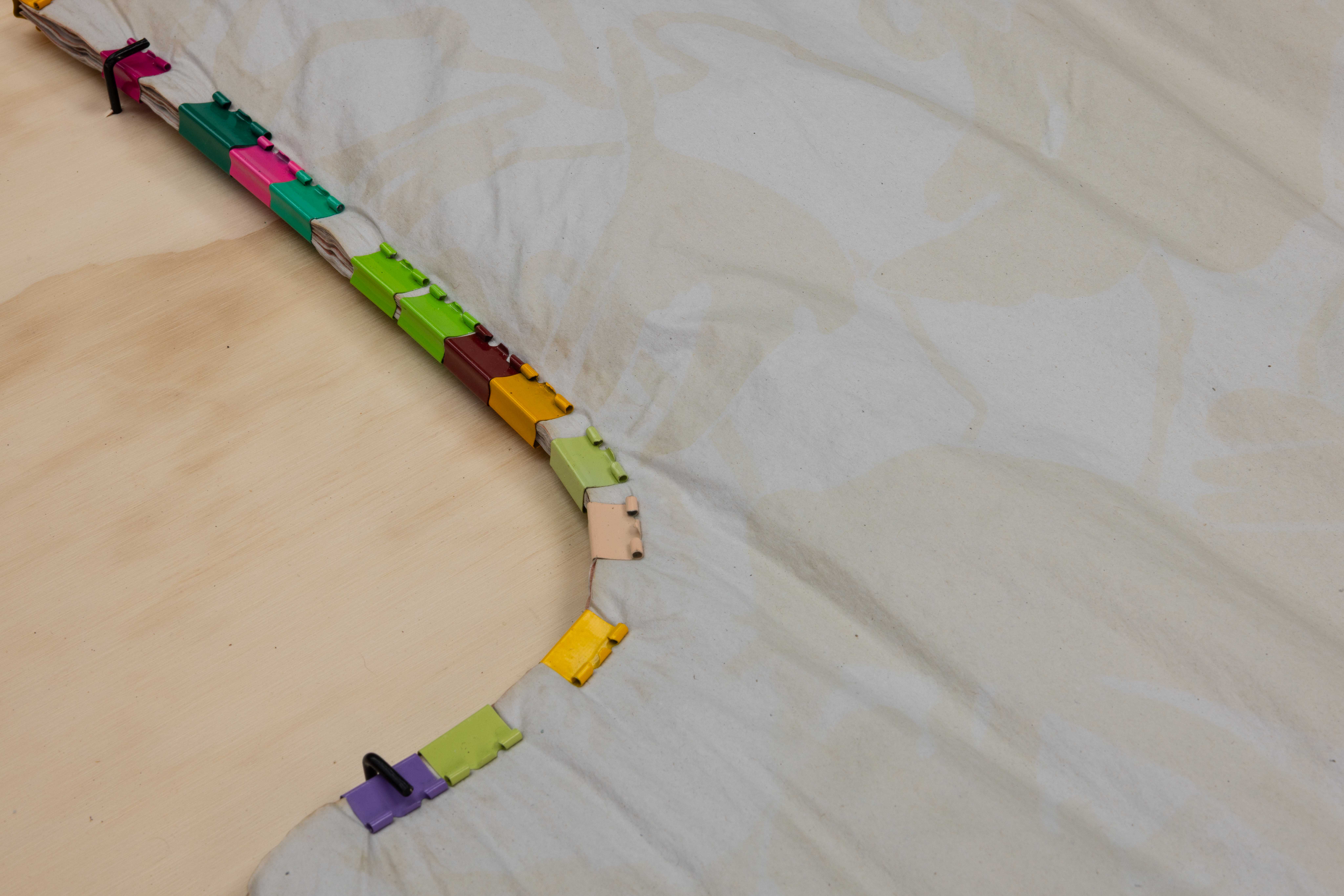 Close-up detail of the stained fabric edge fastened to plywood with a row of colourful plastic clips in pink, green, purple and gold along the curved seam.