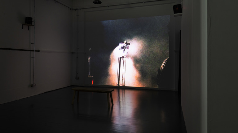 Installation view of a dark gallery space with a wooden bench facing a large video projection. The projected footage shows a grainy, backlit scene of a figure standing on a ladder or scaffold-like structure, silhouetted against bright, hazy light with smoke or steam rising around them. The polished dark floor reflects the glow of the screen.