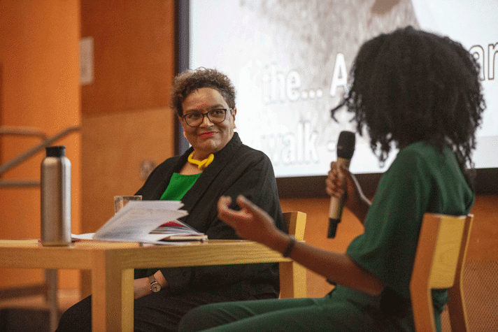 two people sat at a table talking in front of a screen, one holds a microphone