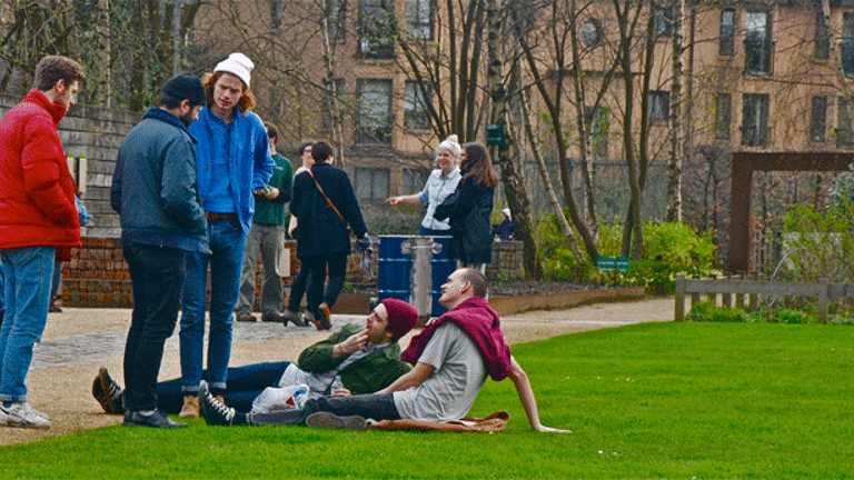 In a garden, people are in the middle of a conversation. Some are sitting on the grass, while some are standing and talking to them. Behind them there is another group of people standing and talking around a couple of trash cans. 