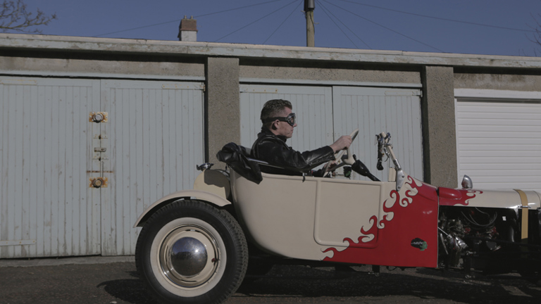 Person sitting in a custom made car in front of some garages 