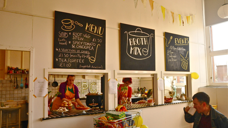 In a cafe, the kitchen is visible through windows made into the wall. There is a customer sipping his coffee and looking towards the kitchen. Through the three windows, two women working in the kitchen are visible.