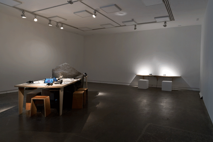Installation view, two illuminated tables with stools displaying small screens and headsets
