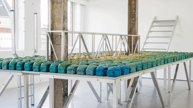   White room with metallic table in the middle, with two vertical cement beams across it. The table is filled with loafs of bread in blue and green tones. 