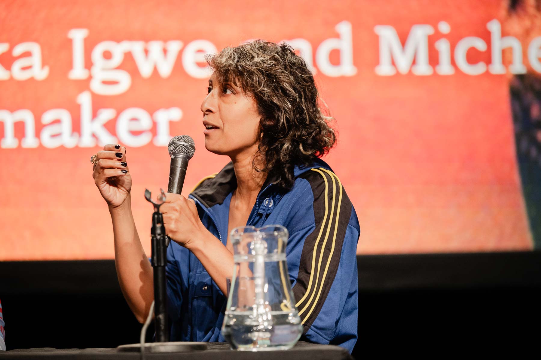 Close-up of a woman with curly hair speaking into a microphone on a panel stage, gesturing with her hand, wearing a blue and yellow striped jacket, with a water jug on the table in front of her.